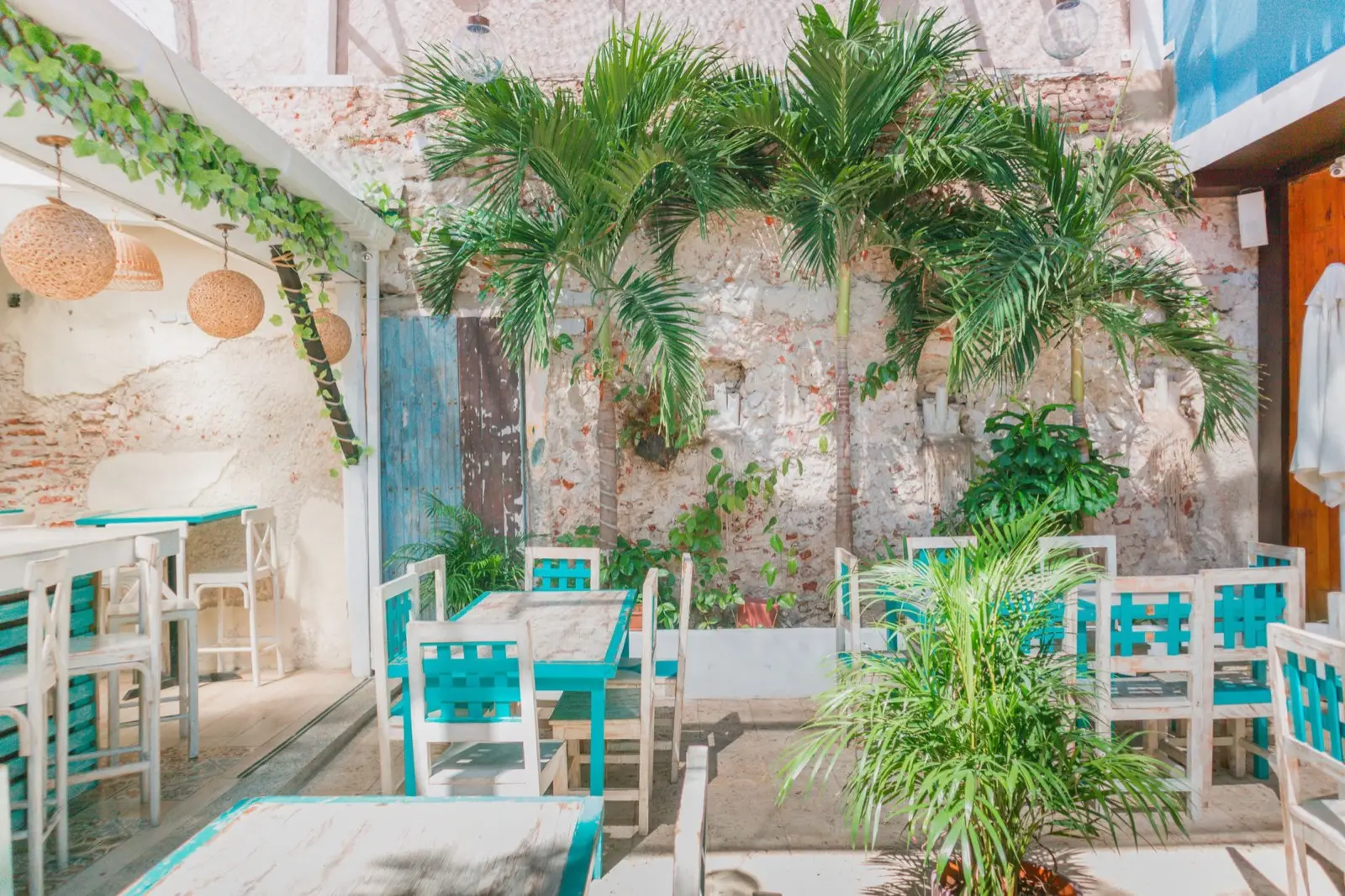Patio con palmeras, muro de piedra y mesas blancas con sillas turquesa